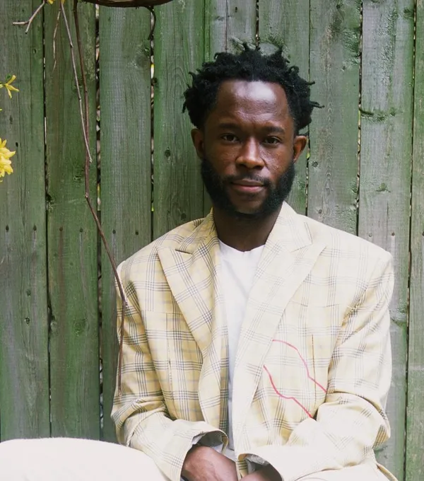 The filmmaker Arie Esiri. A bearded Black man in a white suit sits in front of a pale greyish-turquoise fence.