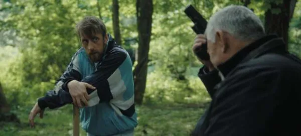 A still from the film Lucky Liam. Two white men stand in deciduous woodland. The younger one is leaning on a spade. The older one is gesticulating with a gun.