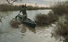 A man pulls a boat out of the reeds, gets into it, and paddles from a narrow creek covered in phragmites towards a larger river.
