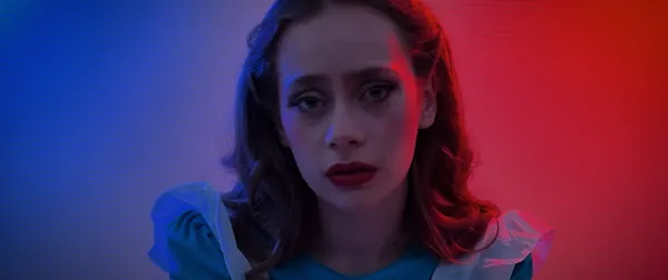 A still from the film The Dark Gets In. The face of a white teenage girl who is dressed in an Alice In Wonderland outfit and lit half in red and half in blue.