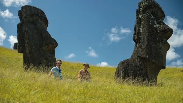 A still from the film Wild Horse Nine. Two white men wander through the long green grass between two of the gigantic stone head statues of Rapa Nui.